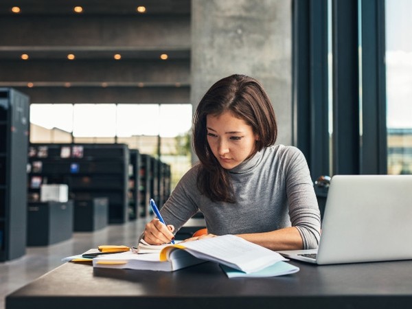 A woman with long dark hair wearing a gray turtleneck is sitting at a table in a modern library, writing in an open book with a blue pen, with a laptop and other books on the table.