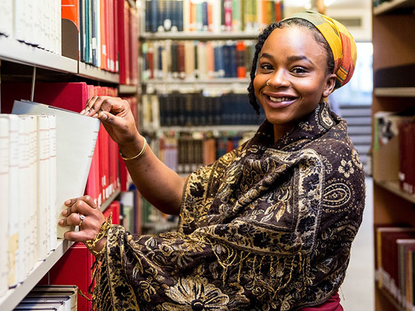 Smiling woman wearing a patterned shawl and a yellow headscarf pulling a book from a shelf in a library aisle.