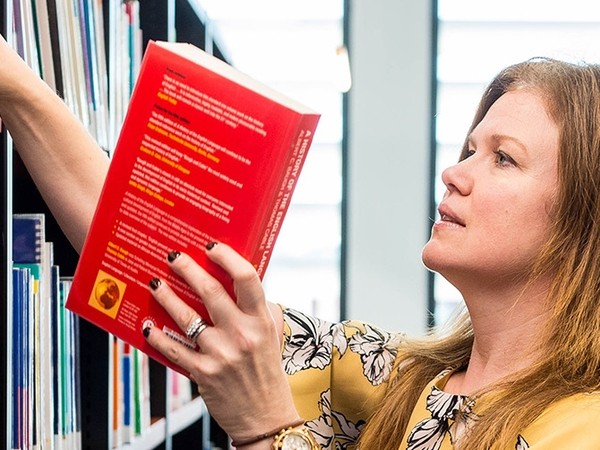 Woman with long hair wearing a yellow floral blouse selecting a red book from a bookshelf in a well-lit room