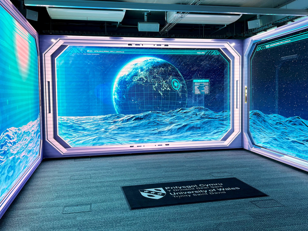 Room with large digital screens on three walls displaying a 3D animated ocean surface and a glowing Earth-like planet with digital interface elements, with a University of Wales Trinity Saint David floor mat centered on the carpeted floor.