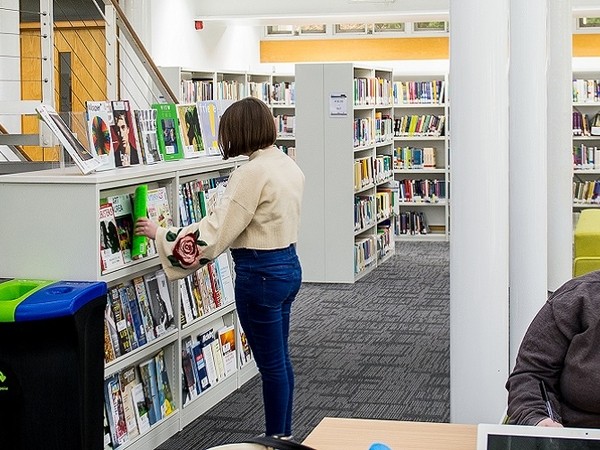 A woman in a cream jacket and blue jeans browsing books on low shelves in a modern library with rows of bookshelves and a carpeted floor.