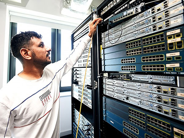 A man in a white Puma Sport shirt working on a server rack filled with networking equipment and cables in a bright room with large windows.
