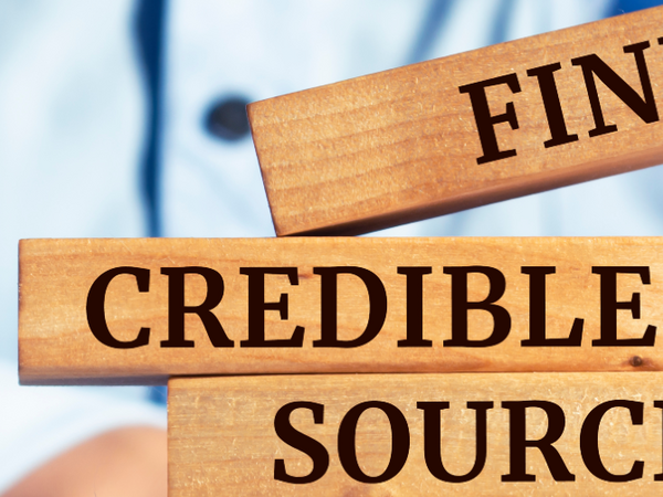 Close-up of wooden blocks with the words FINANCE, CREDIBLE, and SOURCE printed in black, held by a person wearing a blue shirt in the background.