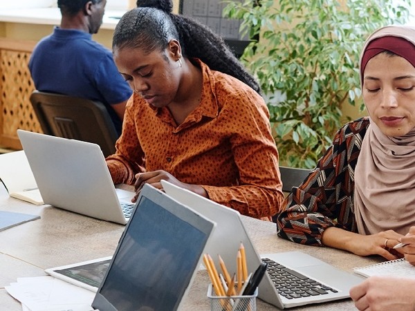 Two women working on laptops at a table, with one woman wearing a rust-colored shirt and the other wearing a patterned hijab, and a man sitting in the background facing away