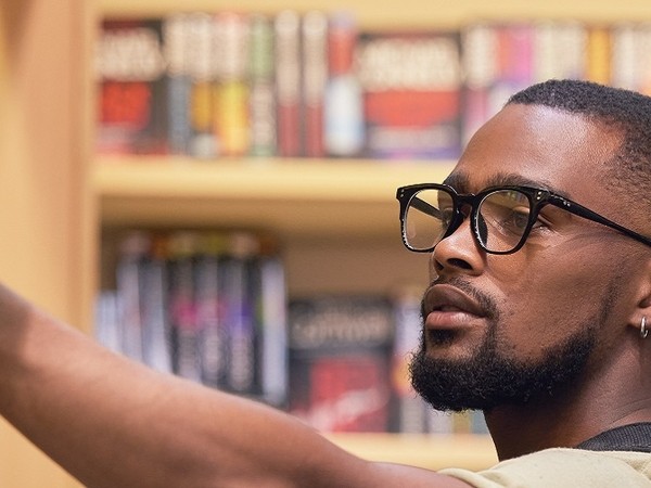 Close-up of a man with glasses and a beard reaching out with his arm in front of a bookshelf filled with books.