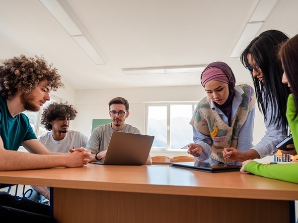 A group of five young adults, including a woman wearing a hijab and a woman pointing at a tablet, gathered around a table in a bright room with a laptop and tablet, collaborating or studying together.