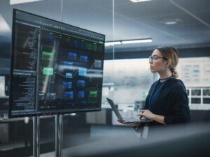 A woman wearing glasses and a dark sweater stands in a modern office holding a laptop, looking at a large monitor displaying complex software code and diagrams.