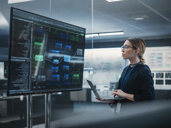 A woman wearing glasses and a dark sweater stands in a modern office holding a laptop, looking at a large monitor displaying complex software code and diagrams.