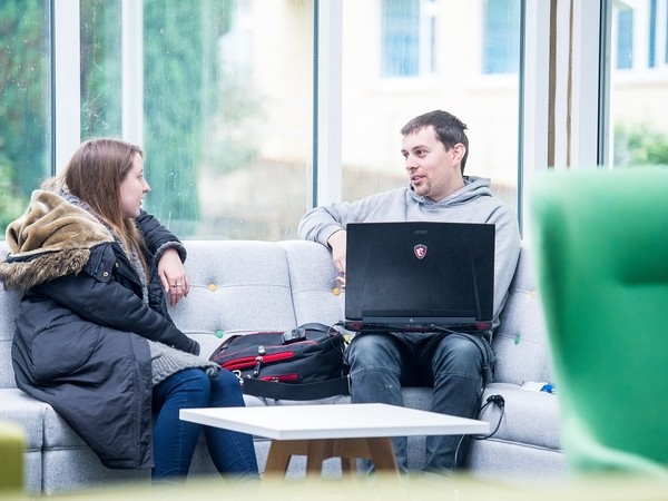 Two people sitting on a light gray couch near large windows, engaged in conversation, with a man holding a laptop on his lap and a woman wearing a black coat with a fur-lined hood.