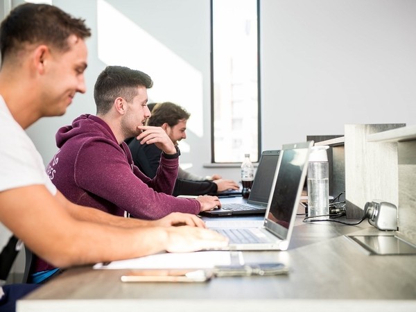 Three men sitting in a row at a desk working on laptops, with a water bottle and other items on the desk and a bright window in the background