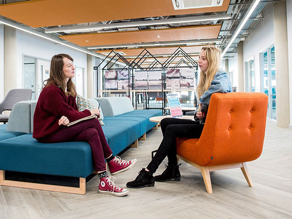 Two women having a conversation seated in a modern office lounge area with one on a blue sofa and the other on an orange armchair, light wood flooring, and shelves in the background.