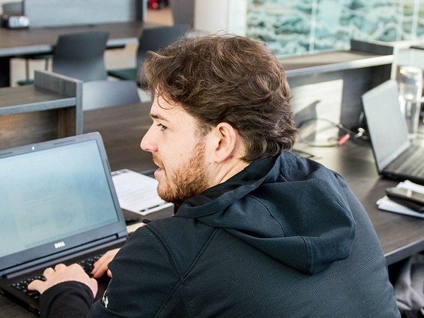 Man with brown hair and beard wearing a black hoodie, sitting at a desk and typing on a laptop in an office environment.