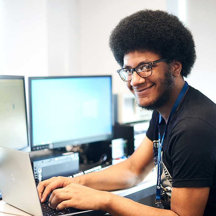A smiling man with glasses and an afro hairstyle working on a laptop at a desk with two computer monitors in the background.
