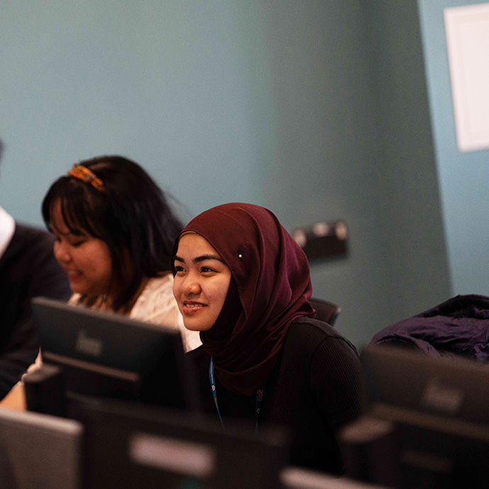 Two women sitting at desks with computer monitors, one wearing a maroon hijab and black top, smiling and looking to the side, the other with dark hair and a headband, in an office or classroom setting.