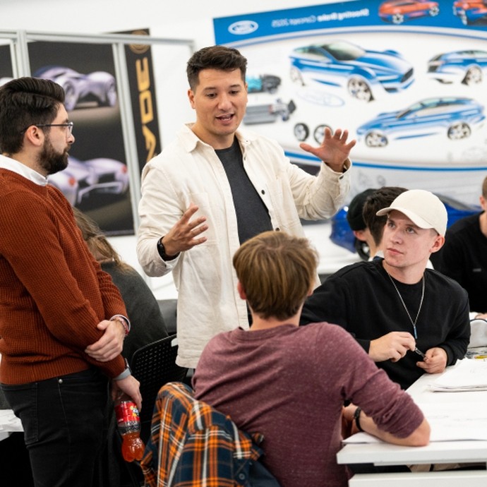 A group of young adults engaged in a discussion, with one man in a white jacket speaking animatedly while others listen, in a room decorated with car design posters and sketches.