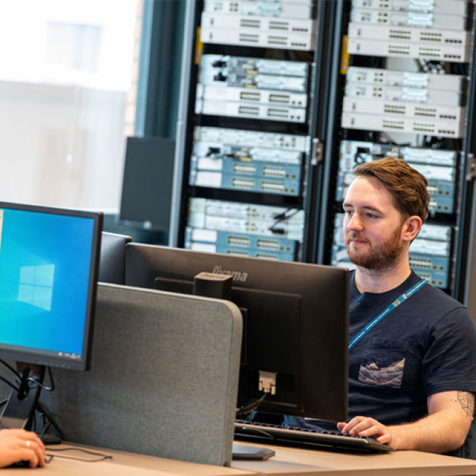 Man with a beard and short brown hair working at a computer desk with multiple monitors in a server room environment.