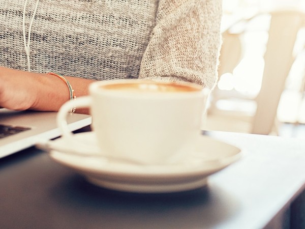 Close-up of a white cup of coffee with a saucer on a table, a person in a gray textured sweater using a laptop in the background