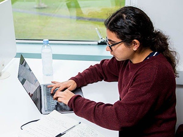 Person with dark hair and glasses wearing a burgundy sweater typing on a laptop at a desk with a notebook, pen, and water bottle near a window.