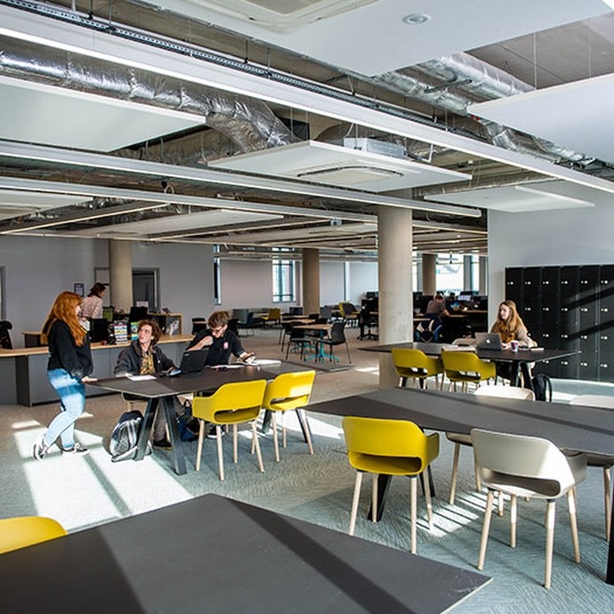 Modern open office space with several tables and yellow and gray chairs, people working and conversing, and industrial-style exposed ductwork on the ceiling.