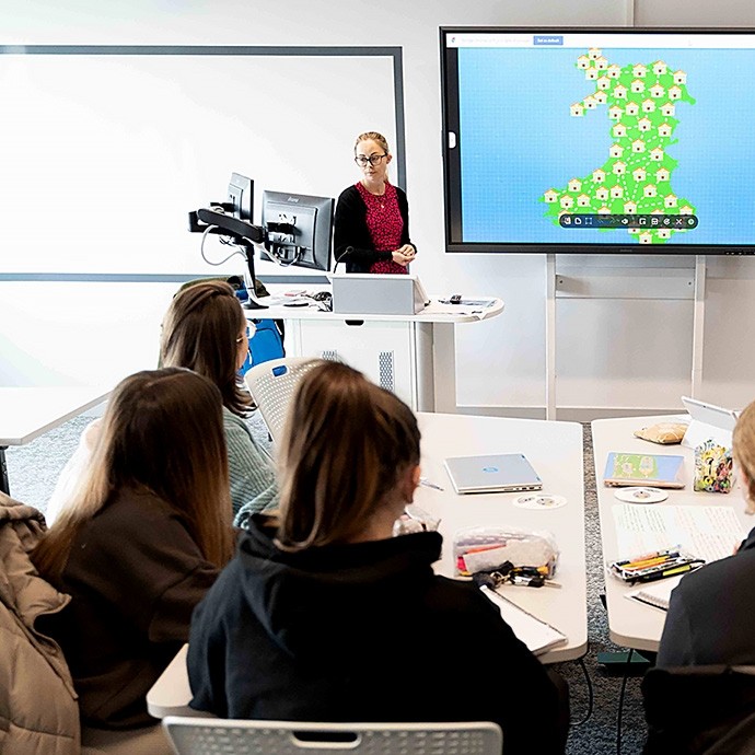 A woman stands at the front of a classroom presenting to a group of students seated at desks, with a large screen displaying a map featuring several icons behind her.