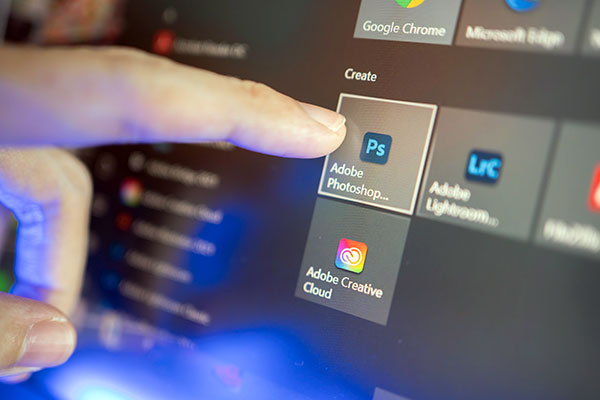 Close-up of a finger pointing to the Adobe Photoshop icon on a computer screen alongside Adobe Creative Cloud and Adobe Lightroom icons with other application icons partially visible.