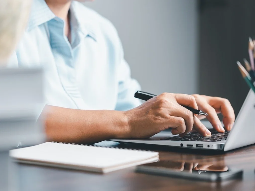 Person wearing light blue shirt typing on a laptop with a pen in one hand, a notebook and a smartphone placed on the wooden table nearby.