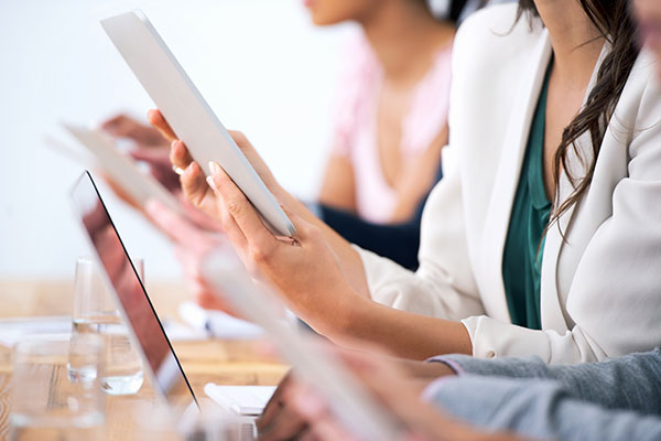 Close-up of multiple people sitting in a row holding digital tablets, with a laptop and glass of water on the table in front of them.