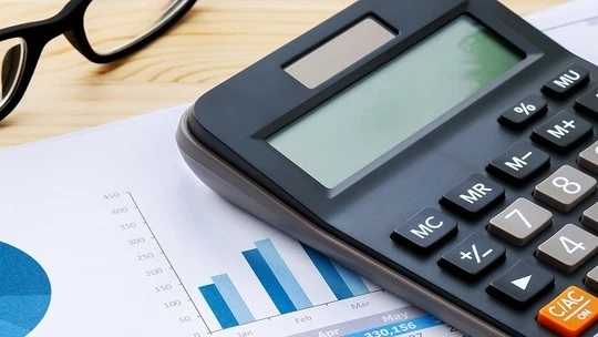 Close-up of a black calculator on top of financial graphs and charts with a pair of black eyeglasses beside them on a wooden surface