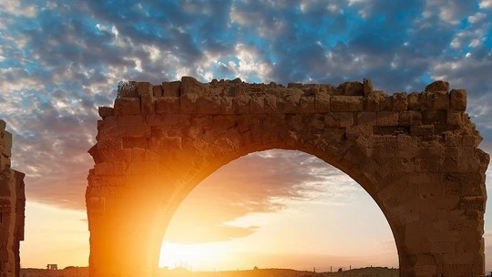 Stone arch ruins silhouetted against a partly cloudy sky during a sunset with warm sunlight glowing through the arch.