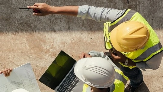 Overhead view of construction workers in safety helmets and vests reviewing plans and a laptop on a concrete surface, with one worker pointing with a pen.