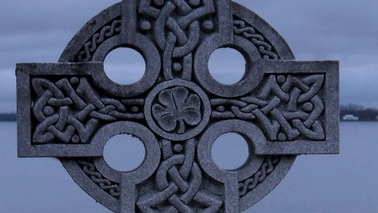 Stone Celtic cross with intricate knotwork design against a background of a body of water and an overcast sky