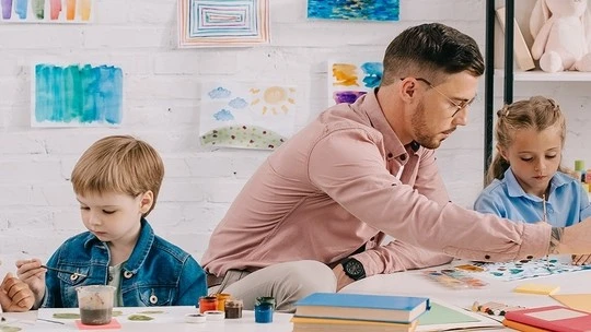 A man and two young children sitting at a table engaged in painting with art supplies and children's artwork displayed on the wall behind them.