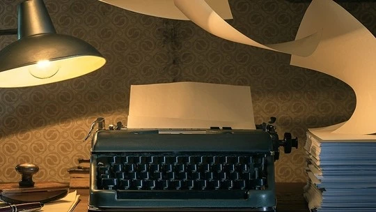 Vintage black typewriter on a wooden desk with a blank sheet of paper loaded, surrounded by stacked papers and illuminated by a hanging lamp with warm yellow light.