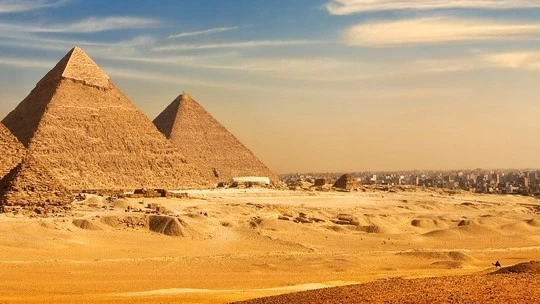The Great Pyramids of Giza under a partly cloudy sky with desert sands in the foreground and a city skyline in the background