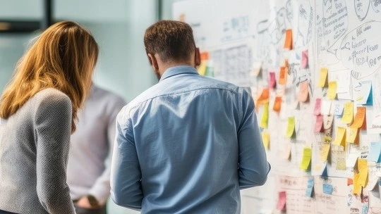 Two people standing in front of a wall covered with colorful sticky notes and papers, appearing to discuss or plan something in a professional setting.
