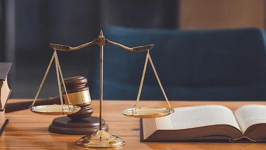 Brass balance scale, wooden gavel, and open book on a wooden desk with a blurred blue chair in the background
