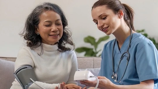 Nurse in blue scrubs measuring blood pressure of smiling older woman sitting on a couch in a home setting.