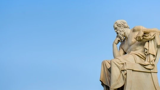 Stone statue of a seated bearded man in classical robes, resting his head on his hand, against a clear blue sky