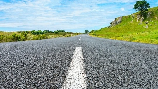 Close-up view of a paved road with a white dashed center line stretching into the distance, flanked by green grassy hills and a partly cloudy blue sky.