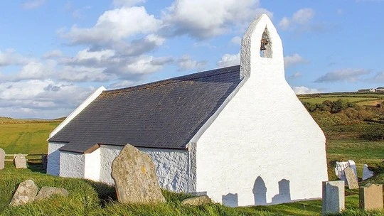 Small white stone church with a single bell tower and slate roof situated in a grassy area with several gravestones and rolling hills under a partly cloudy sky