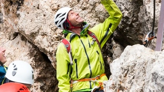 Man wearing a bright green jacket and white climbing helmet looking upwards while rock climbing, surrounded by rocky terrain and accompanied by others in climbing gear.