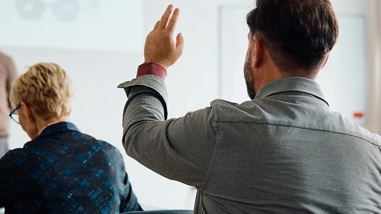 Man raising his hand in a classroom or meeting setting with another person sitting beside him facing forward