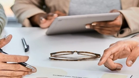 Close-up of hands at a table during a meeting, one holding a pen near a document and another hand touching the table with a pair of eyeglasses in the center, a blurred figure holding a tablet in the background.