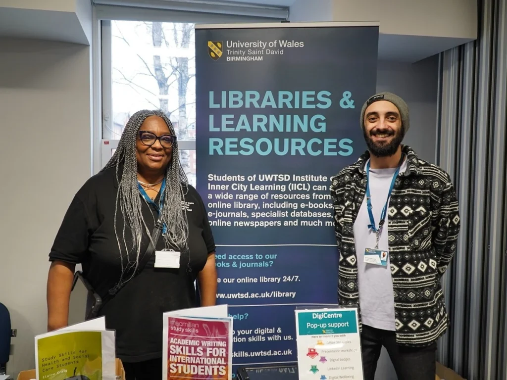 Two people standing indoors in front of a University of Wales Trinity Saint David Birmingham banner that reads Libraries & Learning Resources, with informational pamphlets on a table in front of them.