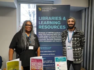 Two people standing indoors in front of a University of Wales Trinity Saint David Birmingham banner that reads Libraries & Learning Resources, with informational pamphlets on a table in front of them.