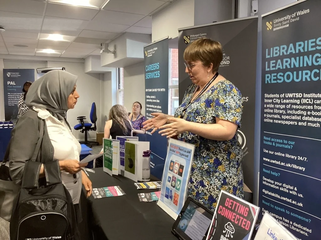 Two women engaged in conversation at a University of Wales information booth with brochures, flyers, and banners about careers services and libraries learning resources in the background.