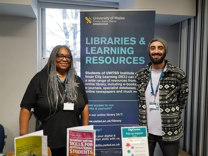 Two people standing behind a table with books and informational materials, in front of a banner for University of Wales Trinity Saint David Birmingham Libraries & Learning Resources, promoting academic writing skills and digital support services
