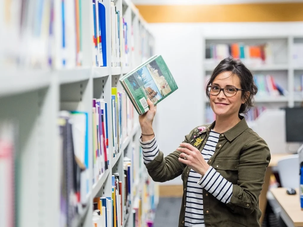 A woman wearing glasses, a green jacket, and a striped shirt holds a book while standing in a library aisle with bookshelves on both sides.