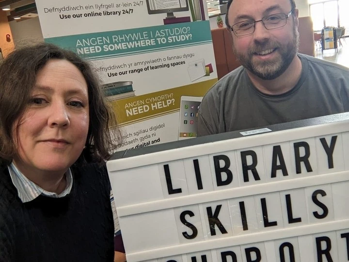 Two people, a woman and a man, smiling and holding a sign that reads 'Library Skills Support' with a background poster offering library study and help services.
