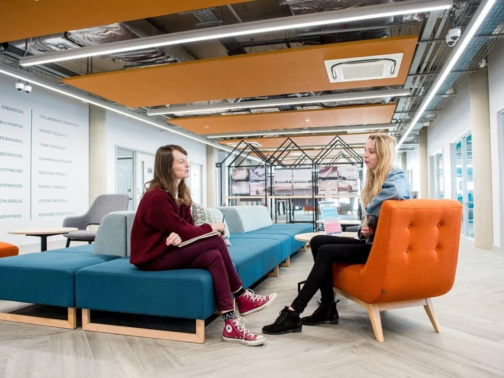 Two women sitting and talking in a modern office lounge area with blue and orange seating, wooden flooring, and exposed ceiling fixtures.